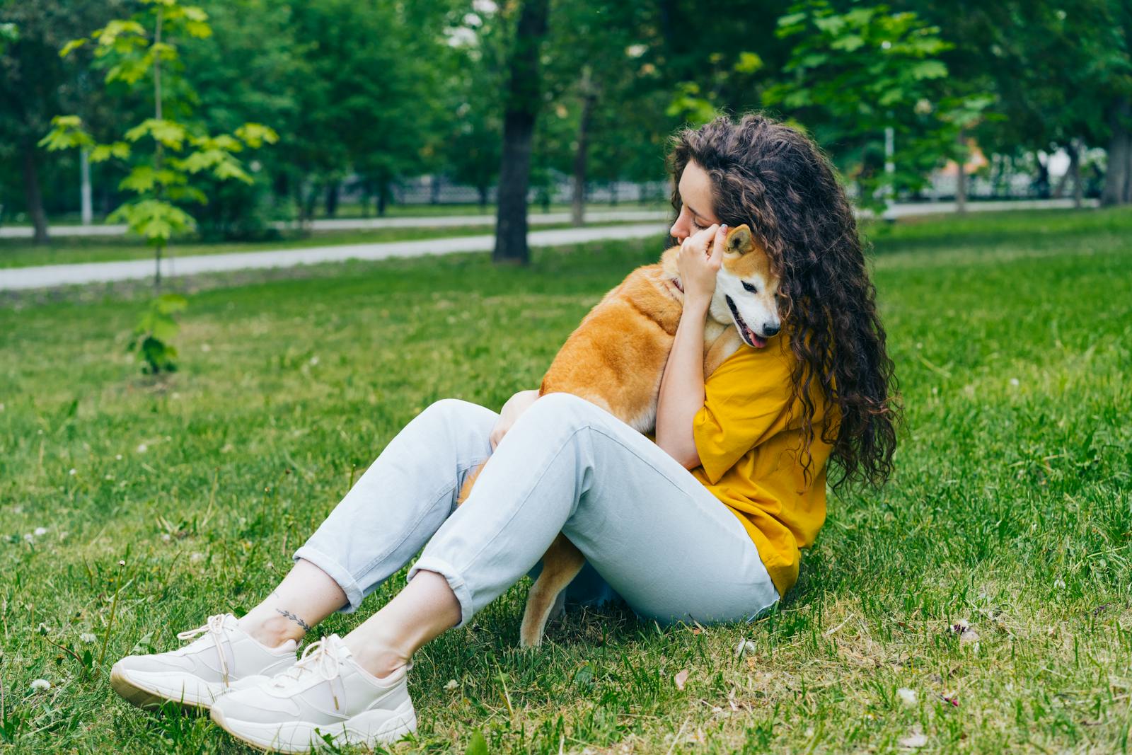 A woman sitting on grass hugging her Shiba Inu dog in a lush green park.