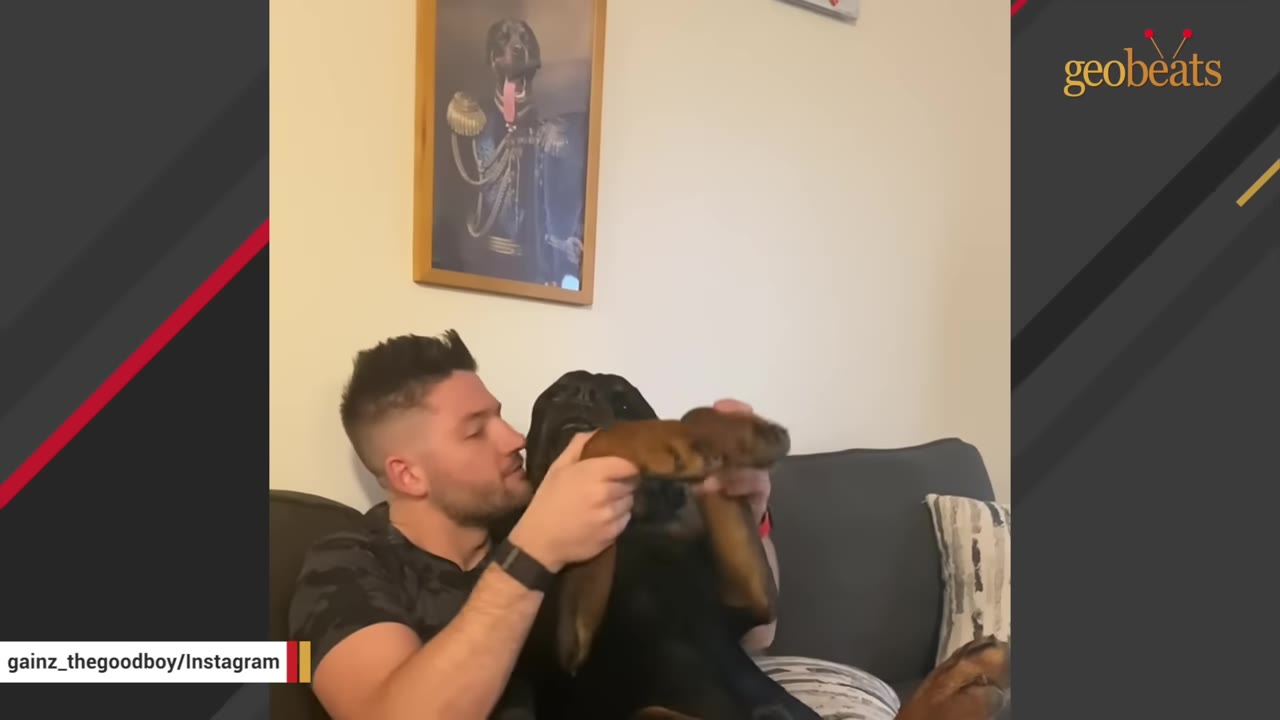 A man sits on a couch cuddling a large black dog, holding its front paws gently.
