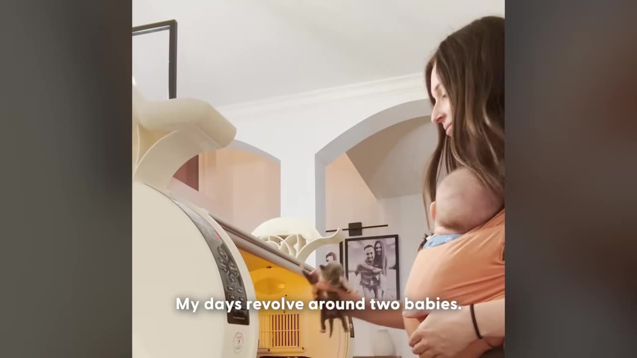 A woman wearing a baby carrier with an infant inside holds a tiny puppy in her hand next to an incubator.