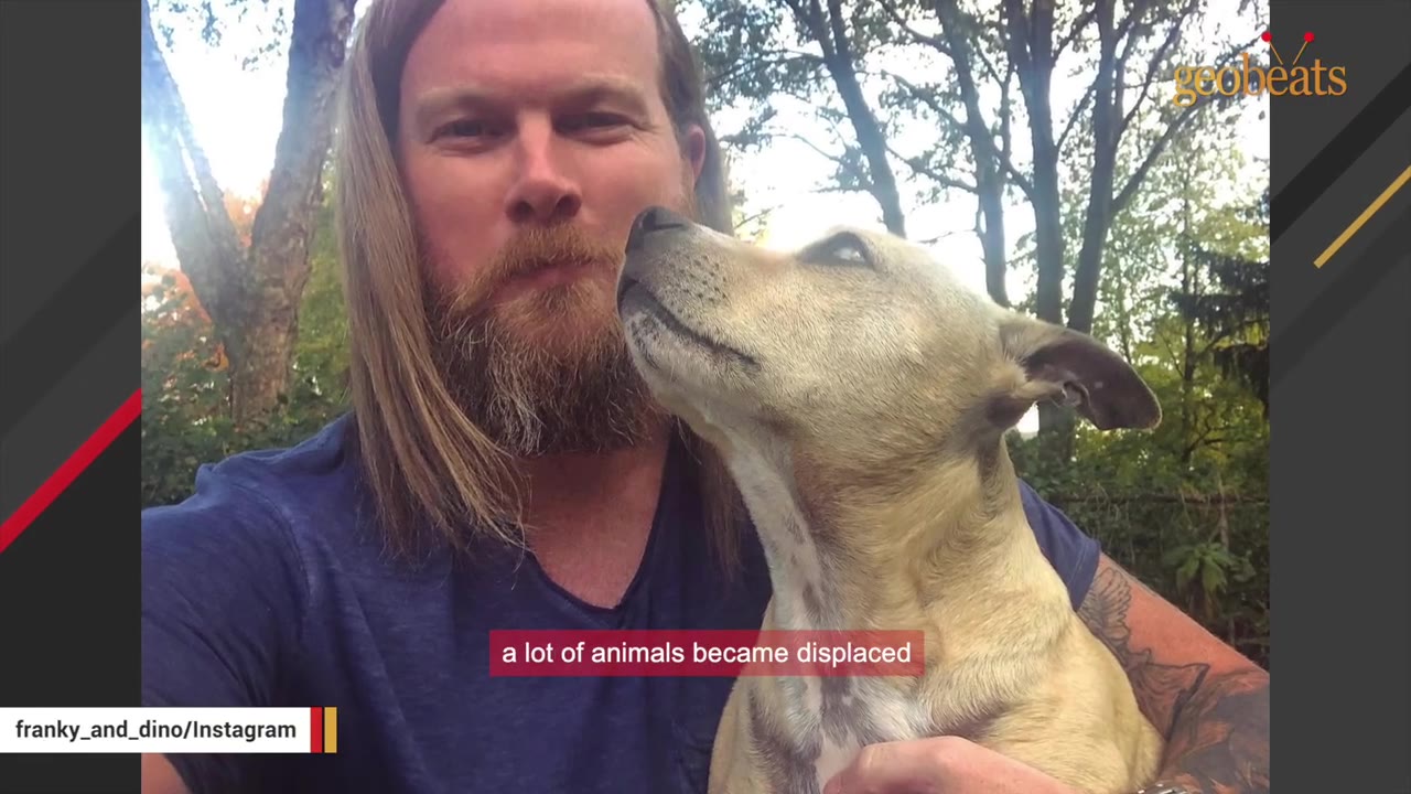 A man with long blonde hair and a beard holds a light-colored dog that is looking up affectionately at him.