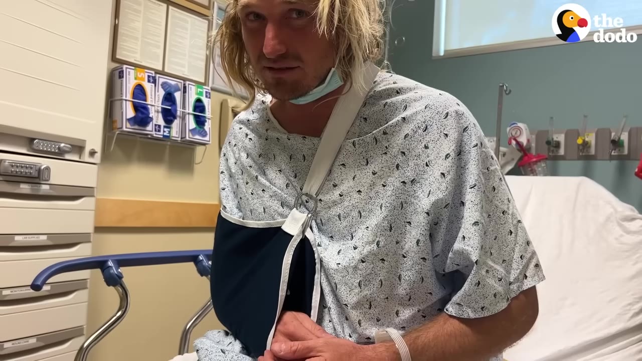 A man with blonde hair wearing a hospital gown sits in a hospital room with his right arm in a black sling.
