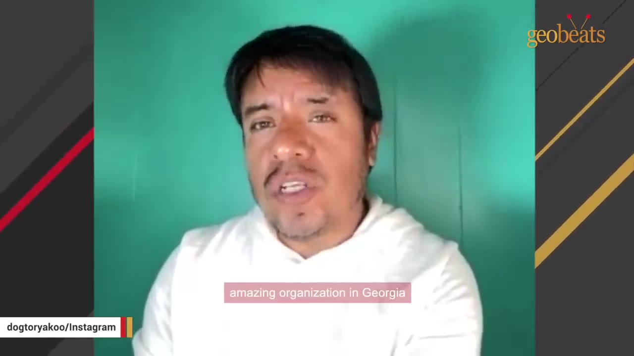 A man with dark hair and a white hoodie speaks in front of a green background.