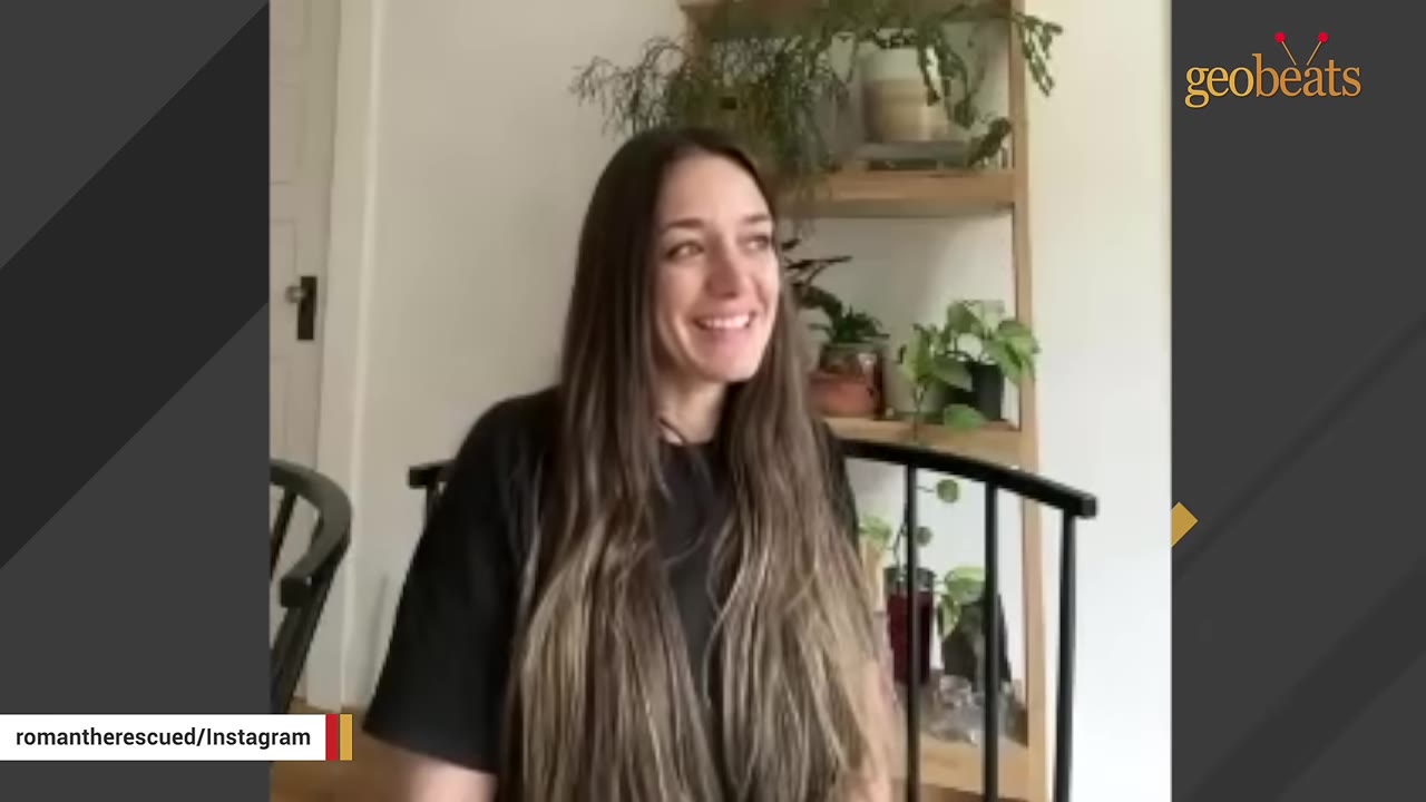 A smiling woman with long brown hair sitting in front of a wooden shelf with potted plants.