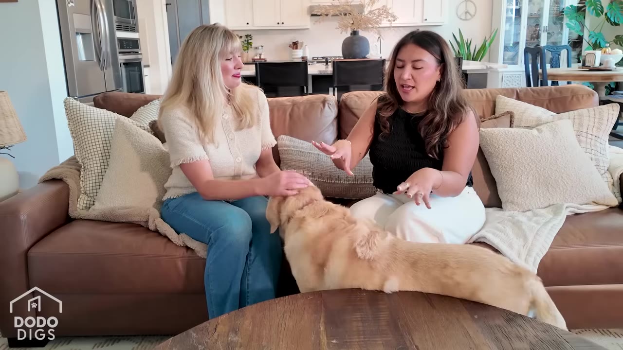 Two women sit on a brown leather couch in a bright living room, smiling and petting a golden retriever dog that stands in front of them.