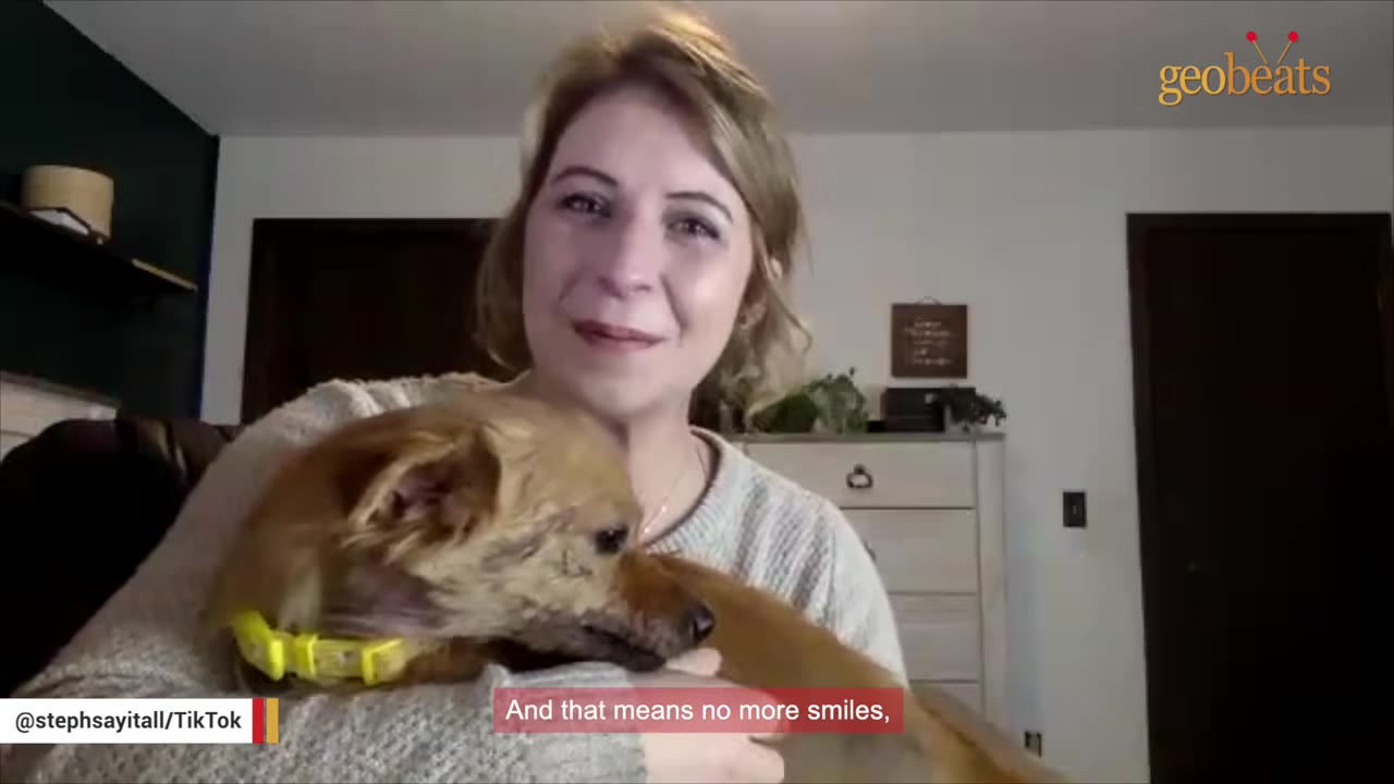 A woman with blonde hair pulled back holds a small brown dog wearing a yellow collar close to her chest, looking emotional.