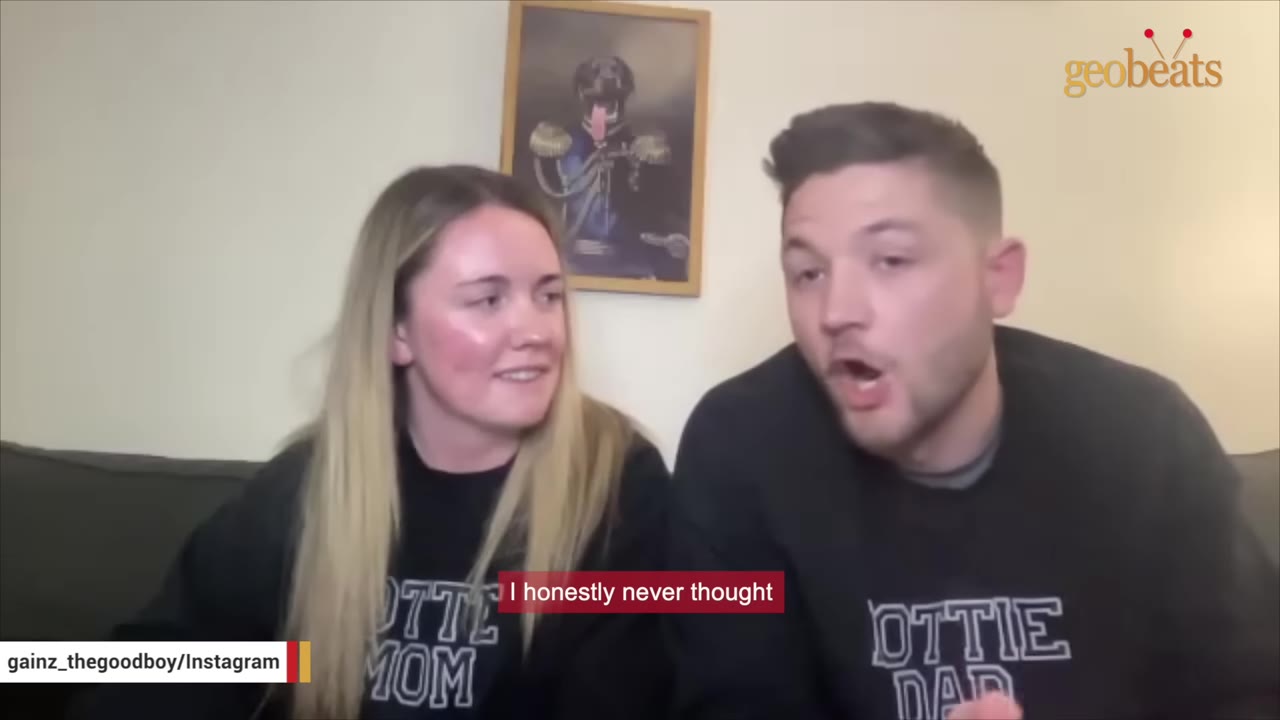 A woman wearing a black sweatshirt that says 'Rottie Mom' looks smilingly at a man wearing a black sweatshirt that says 'Rottie Dad' as he speaks.