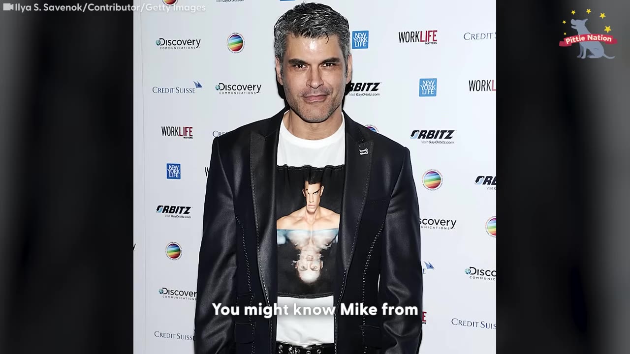 A medium shot of Mike Ruiz posing on a red carpet in a black blazer over a white graphic t-shirt.