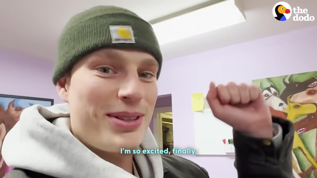 A young man wearing a green beanie and a grey hoodie smiles warmly at the camera, raising his fist in excitement.