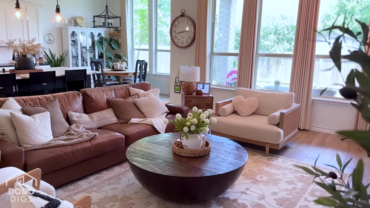 A wide shot of a bright, open-concept living room featuring a large brown leather sectional sofa, a round wooden coffee table, a cream-colored armchair, and a dining area visible in the background.