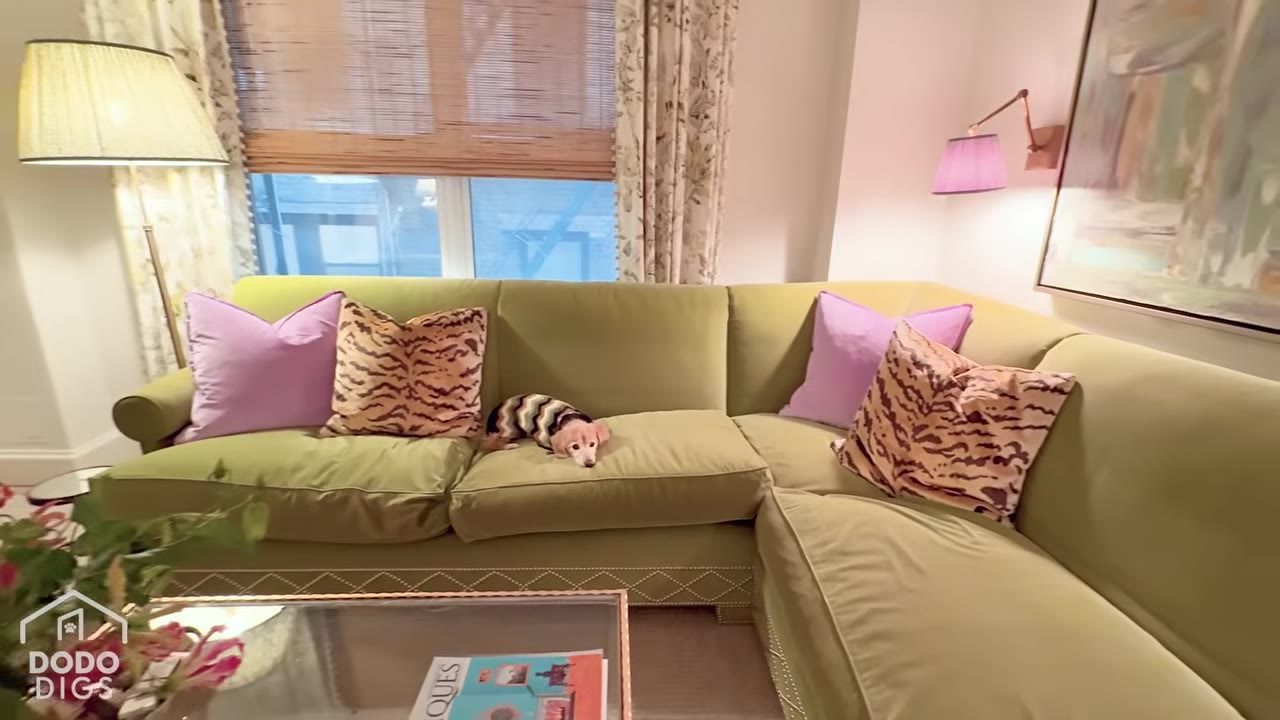 A small dog wearing a patterned sweater rests on a light green sectional sofa adorned with purple and tiger-print throw pillows in a stylishly decorated living room.
