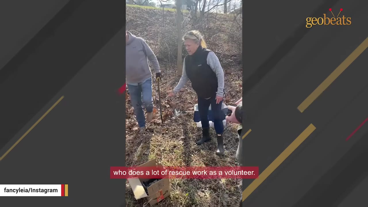 A woman in a black vest and boots stands in a wooded area with dry leaves on the ground, gesturing towards a cardboard box while another person stands nearby.