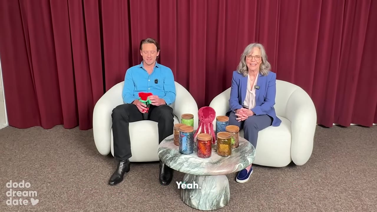 Actors Sally and Lewis sitting in white armchairs in front of a red curtain, with a small marble table in front of them holding several glass jars and a plush octopus toy.