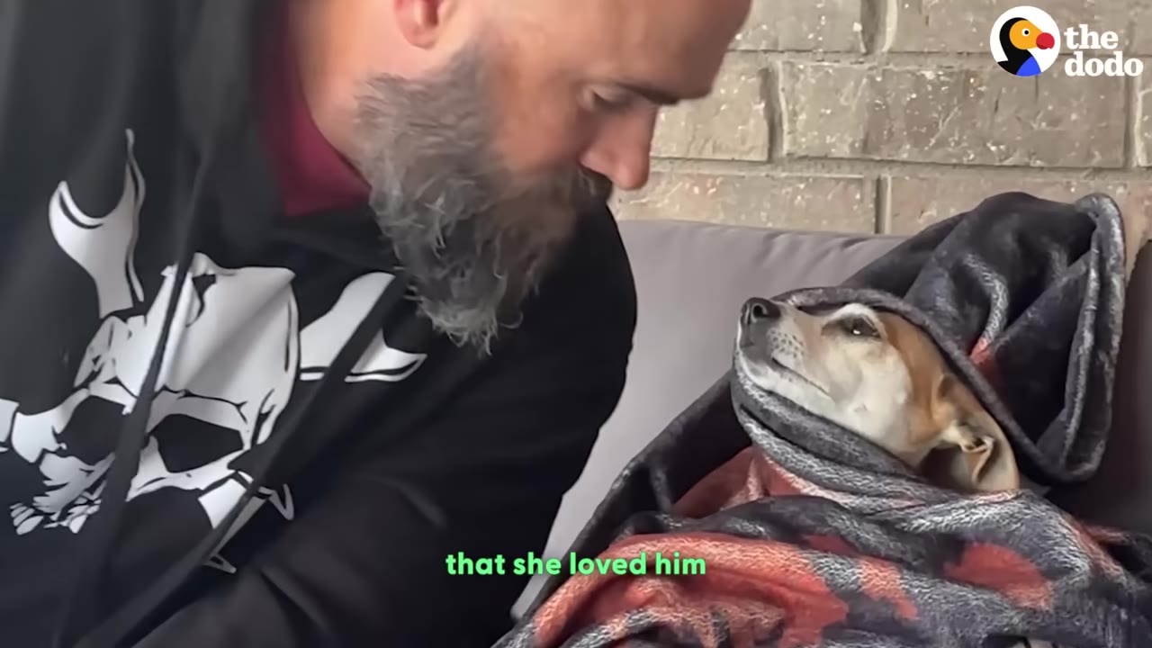 A bearded man looks down affectionately at a small brown and white dog wrapped snugly in a blanket, who gazes back up at him.