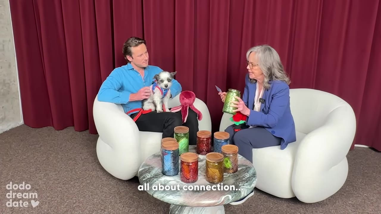 A man in a blue shirt holds a small white and grey dog on his lap while sitting in a white armchair, as a woman in a blue blazer sits opposite him holding a green glass jar and a small card, with several other colorful glass jars arranged on a table between them.