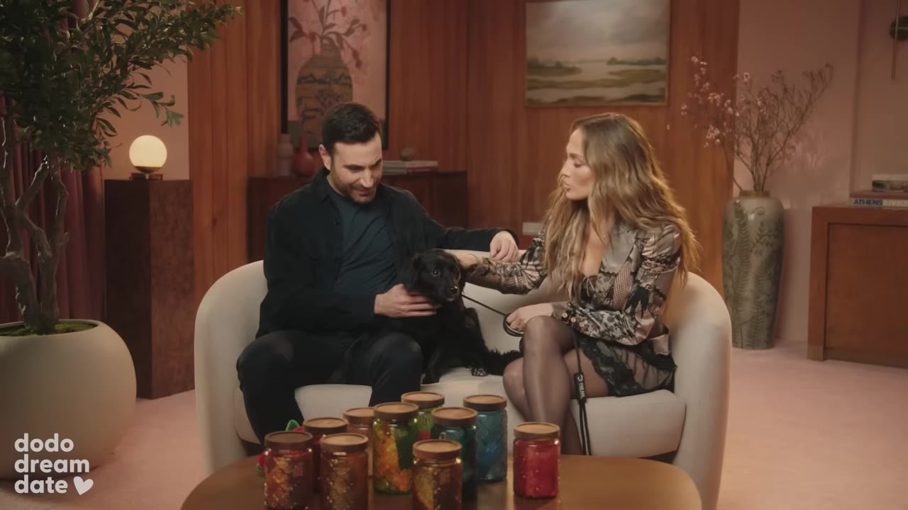 A man and a woman sit on a white couch petting a small black dog, with several colorful jars arranged on a coffee table in front of them.