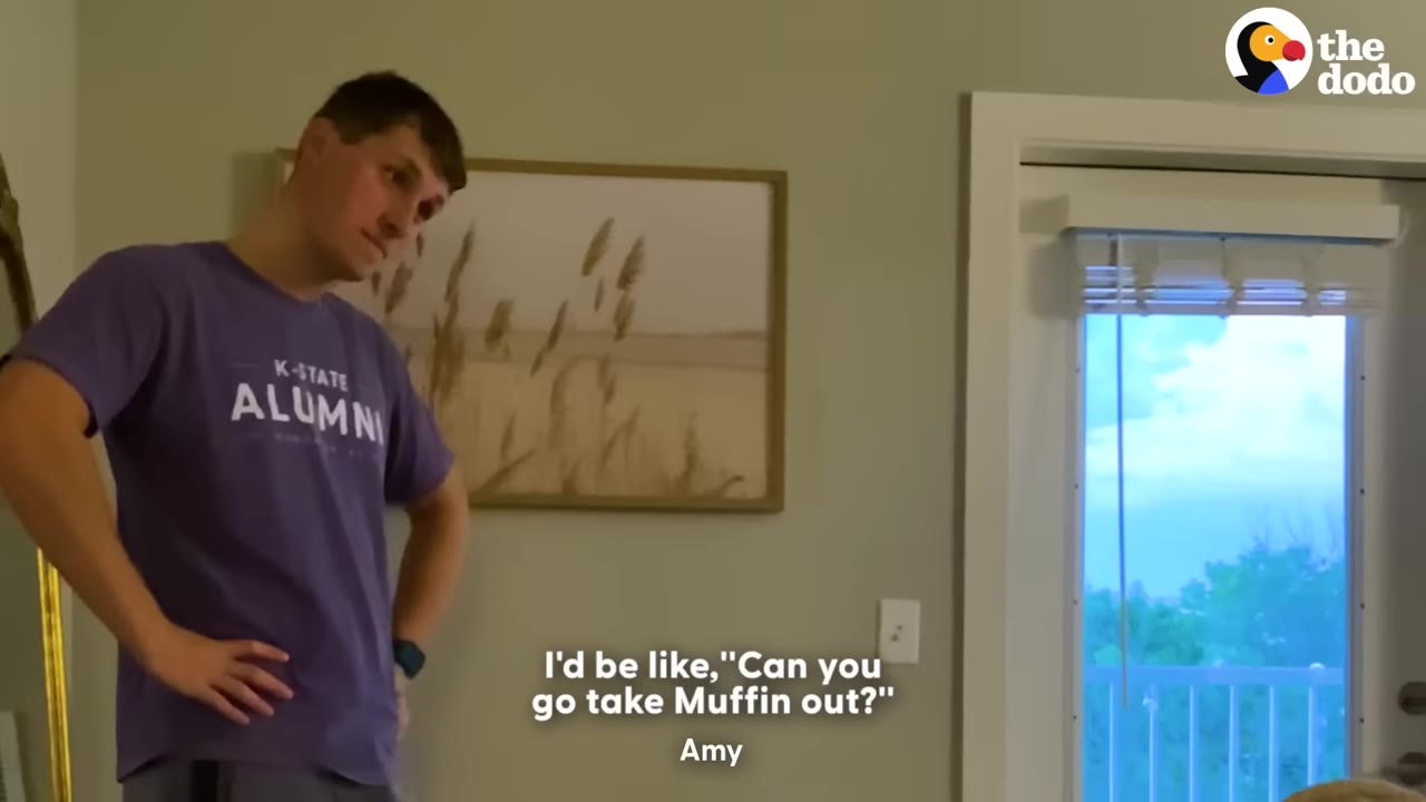 A man in a purple t-shirt stands with his hands on his hips inside an apartment near a glass door.