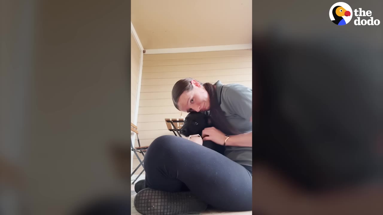 A woman sitting on a porch floor, smiling as she cuddles a small black puppy close to her chest.