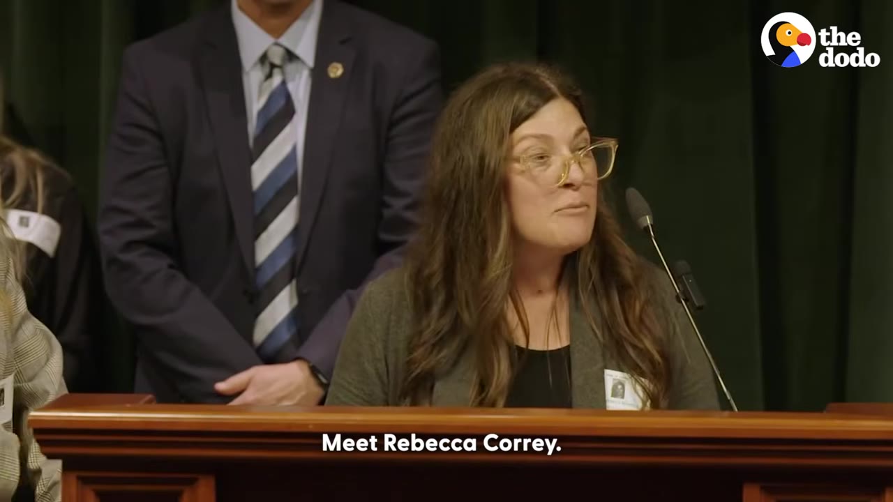 A woman with long brown hair and glasses speaking at a podium with a microphone, with a man in a suit standing behind her.