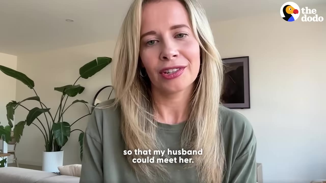 A blonde woman in a green shirt speaks to the camera in her living room, with subtitles reading 'so that my husband could meet her.'