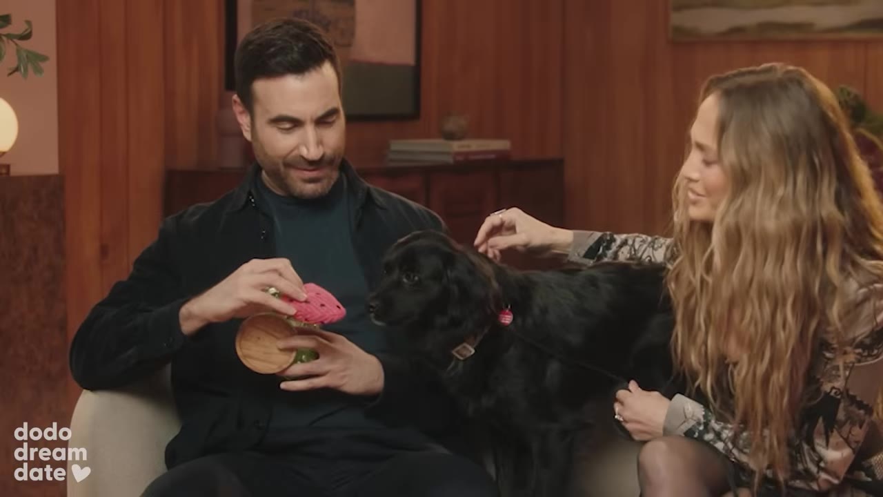 A man holds a pink watermelon-shaped toy in front of a black dog sitting on a couch between him and a woman.