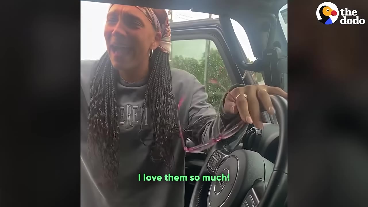 A woman with braided hair and a headscarf looks tearful and emotional as she stands by an open car door, holding her glasses in her hand.