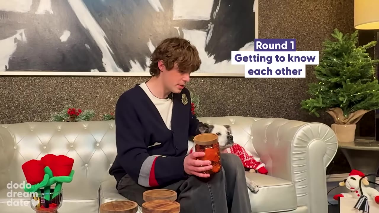 A young man with curly brown hair sits on a silver tufted sofa holding an amber glass jar with a wooden lid, while a small white dog wearing a red and white sweater sits next to him looking away.