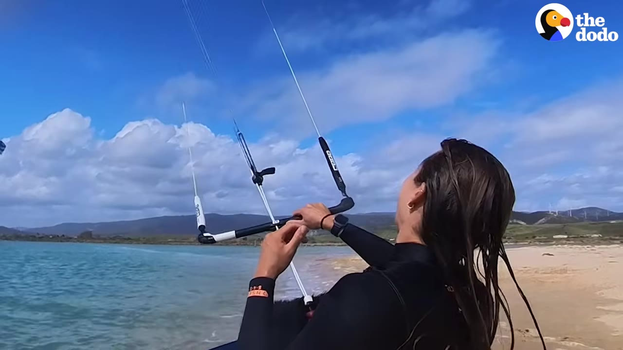 A woman in a black wetsuit stands on a sandy beach, holding onto the control bar of a kitesurfing rig as she looks up at the blue sky.