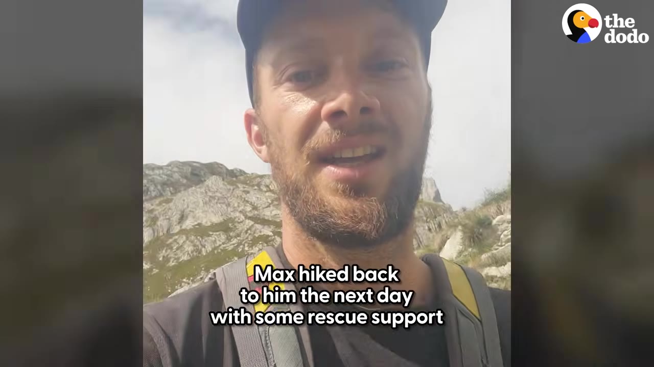 A close-up selfie video frame of a bearded man wearing a cap and backpack, speaking to the camera with a rocky mountain in the background.
