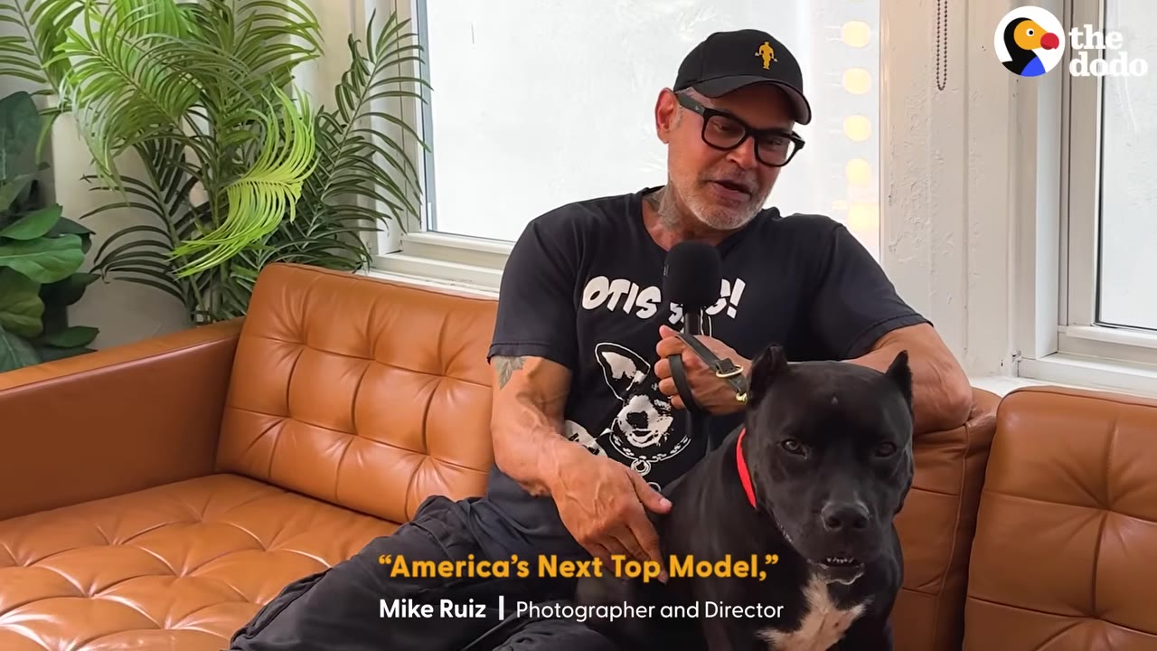 Mike Ruiz, wearing a black cap, glasses, and a black t-shirt, sits on a brown leather couch next to a black dog while holding a microphone.