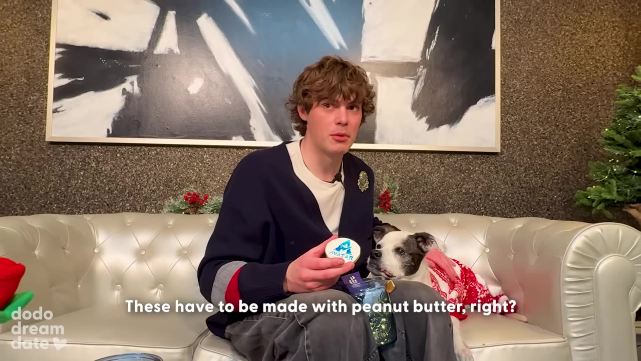 A young man with curly brown hair sits on a white leather sofa holding a round dog cookie with an Avatar logo, while a black and white dog wearing a red and white sweater sits next to him.