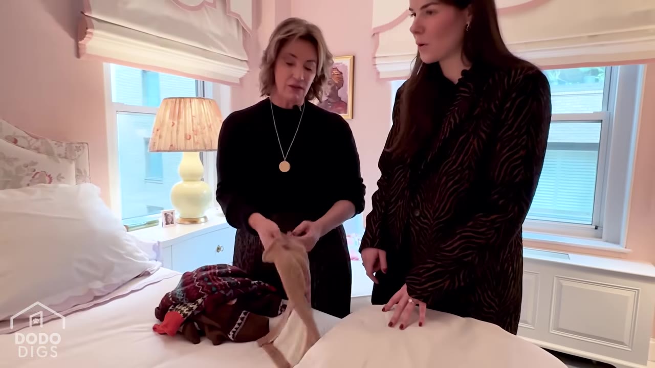 Two women stand by a bed in a pink bedroom, looking at a small tan dog sweater that one of them is holding up from a pile of sweaters.