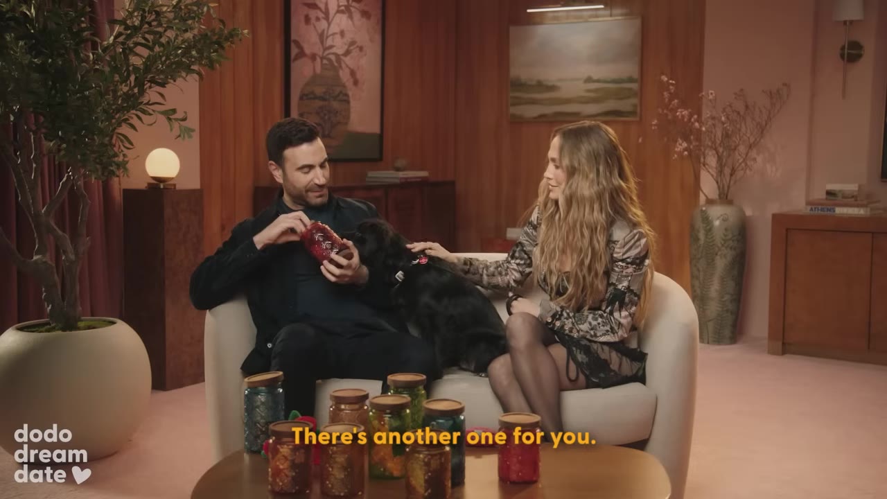 A man sitting on a couch holds a jar of treats for a small black dog, while a woman sits next to them and pets the dog.