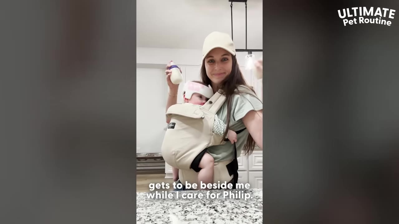 A woman wearing a baseball cap and a baby carrier with a baby inside holds up a baby bottle in a kitchen.