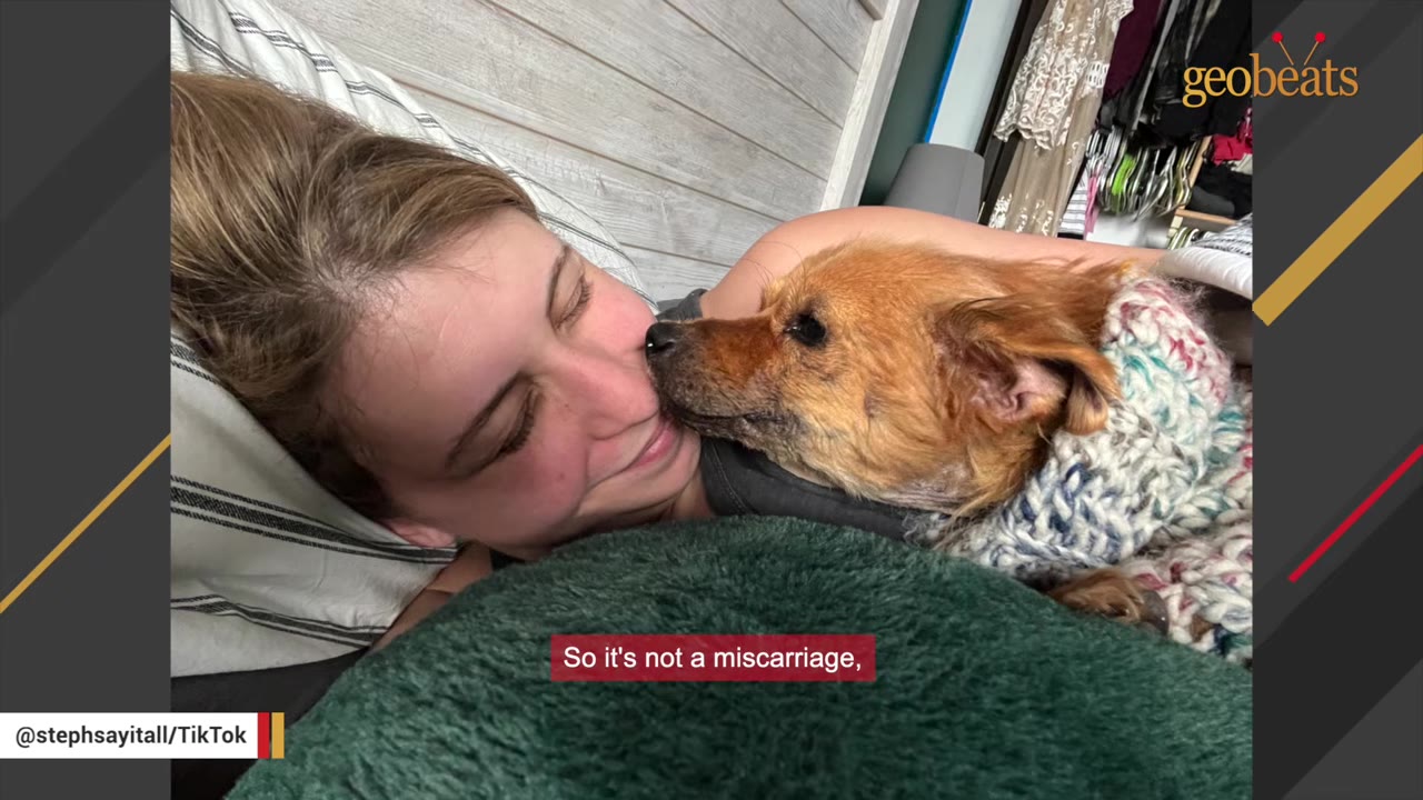 A woman lies on a pillow smiling as a small brown dog wearing a knit sweater nuzzles her cheek.