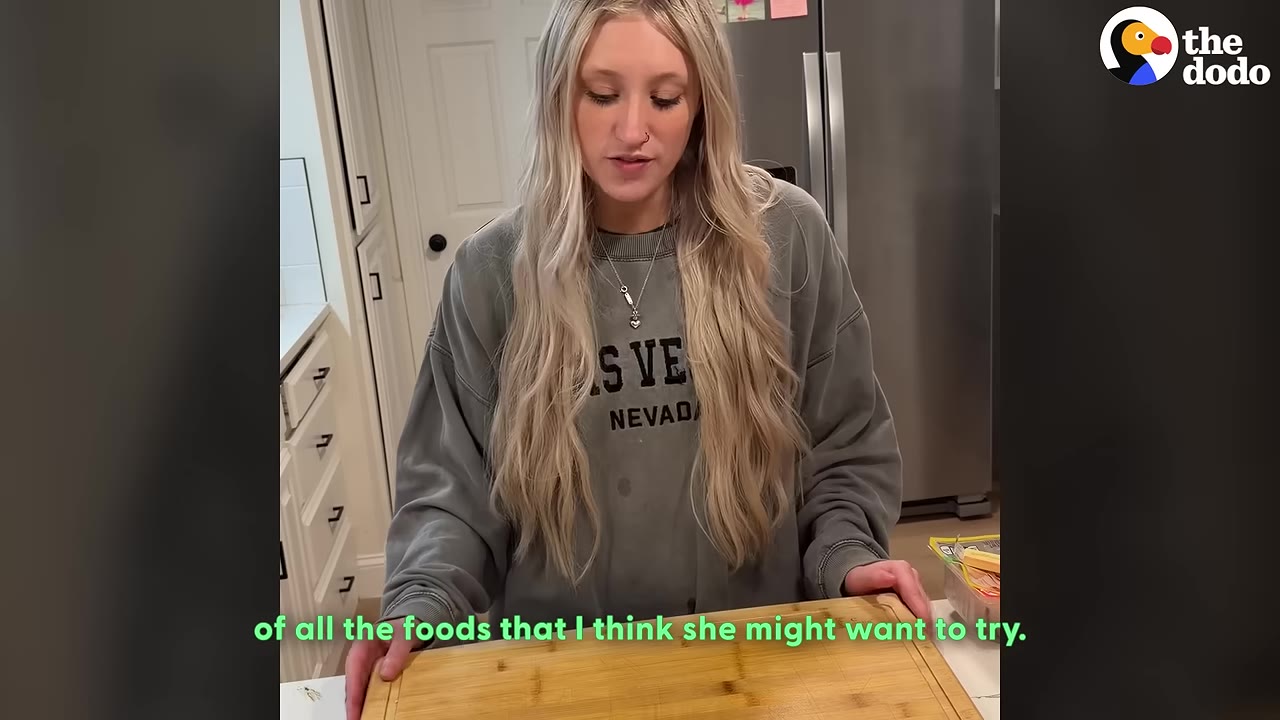 A woman with long blonde hair wearing a grey sweatshirt stands in a kitchen behind a wooden cutting board, looking down.