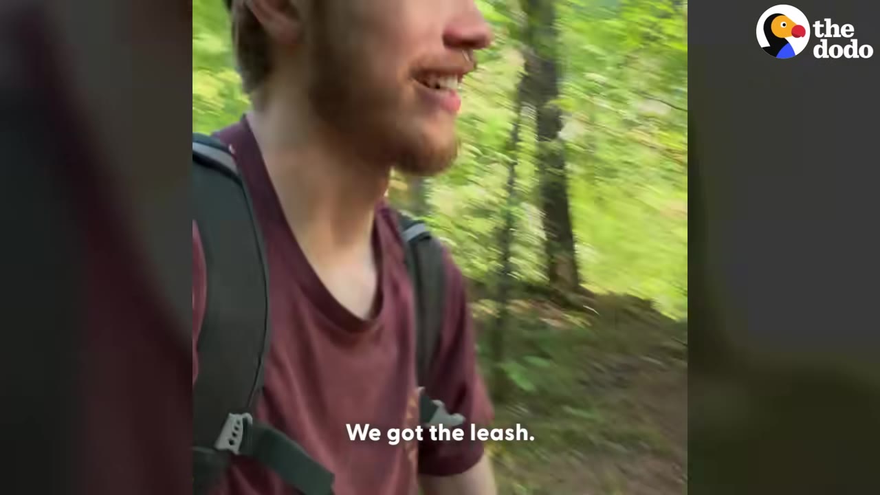 A close-up of a smiling hiker in a maroon shirt and backpack walking through a wooded area, with the text overlay 'We got the leash.'