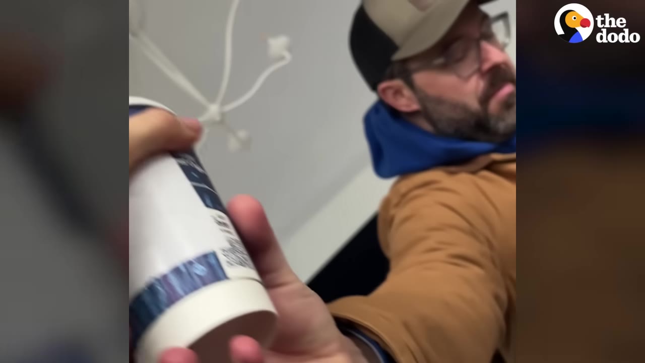 A man wearing a cap, glasses, and a brown jacket hands a white paper coffee cup toward the camera.