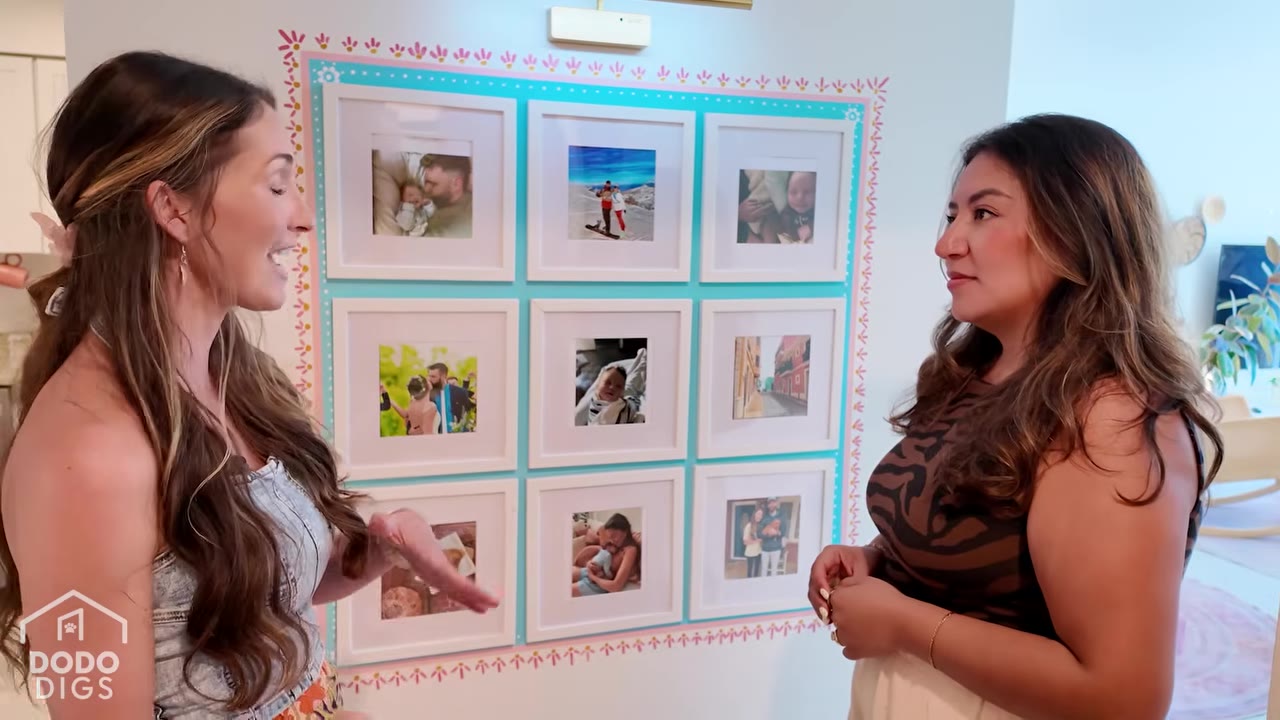 Two women stand in front of a wall gallery of framed family photos, with one gesturing toward the pictures as they talk.