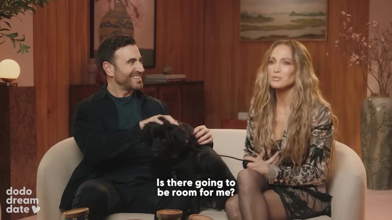 Jennifer Lopez and Brett Goldstein sit on a couch with a small black dog, smiling and talking during an interview.