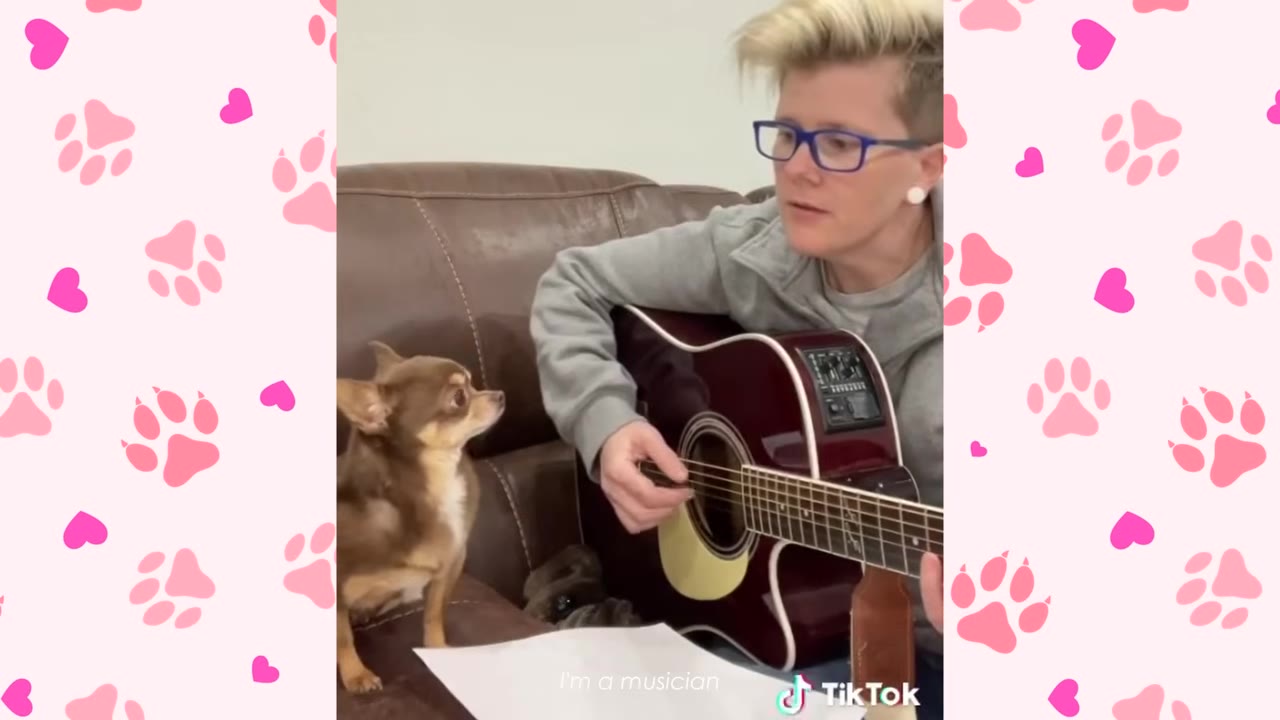 A person with short blonde hair and blue glasses sits on a brown leather couch playing an acoustic guitar while a small brown chihuahua looks up at them.