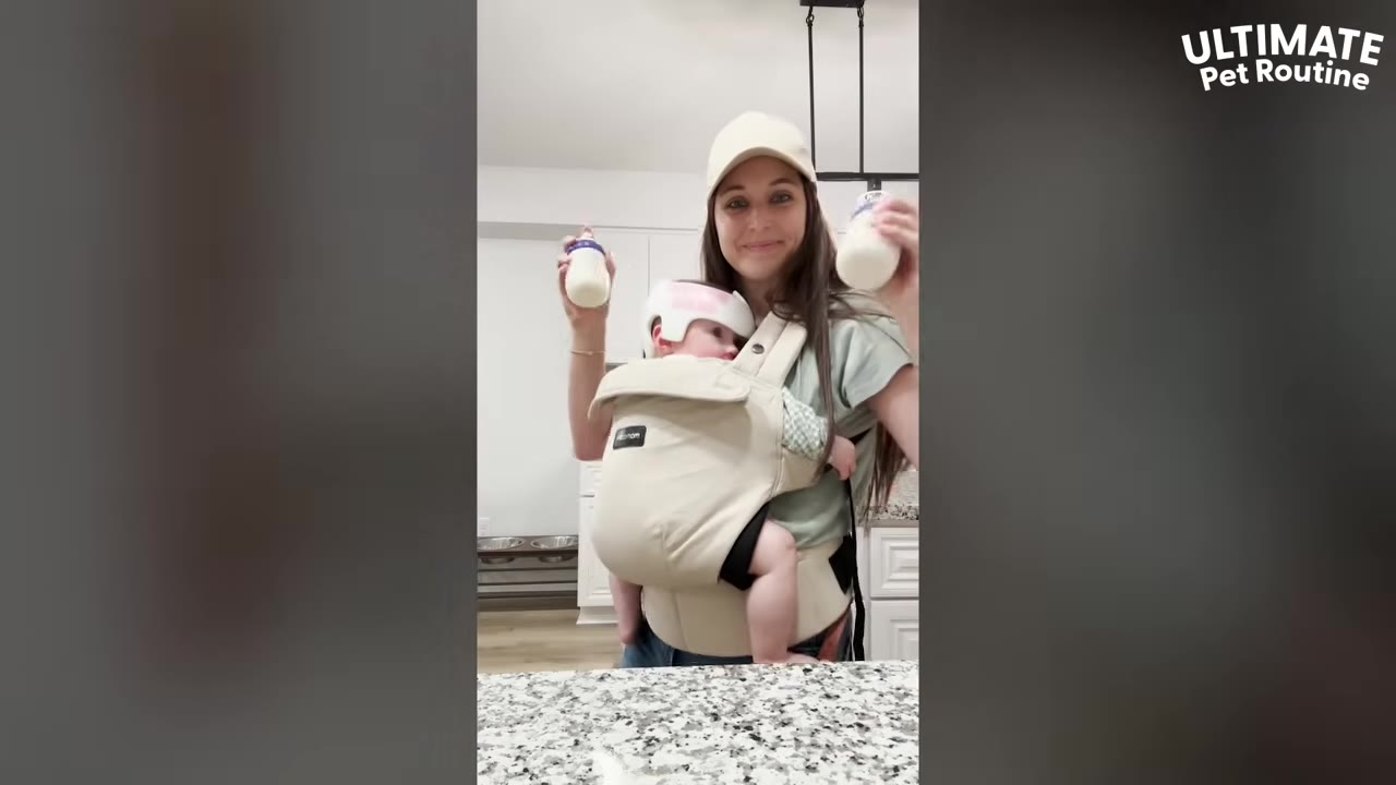 A woman wearing a beige baseball cap smiles at the camera while carrying a baby in a beige front carrier, holding up two baby bottles.