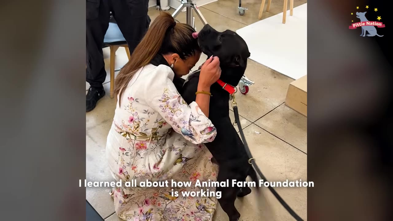 A woman in a floral dress kneels down to hug a black dog wearing a red collar, who is licking her forehead.