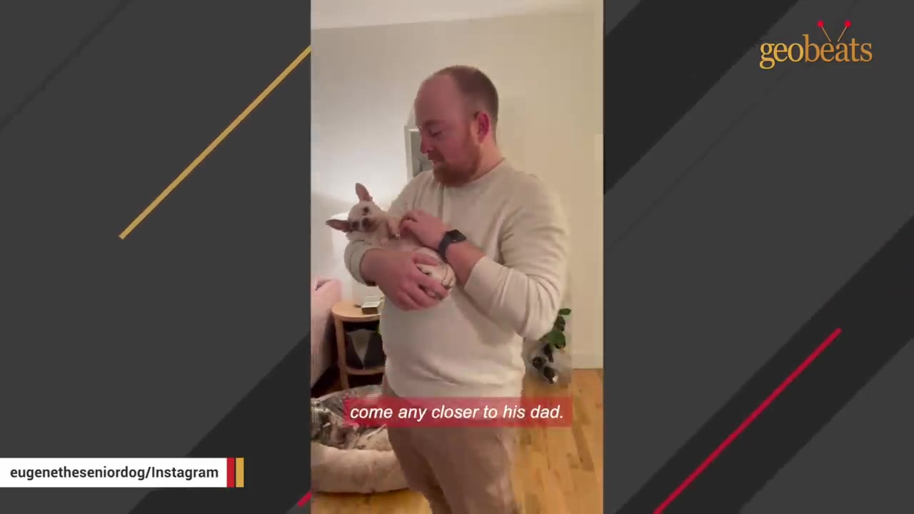 A man holding a small tan chihuahua close to his chest while standing in a living room.