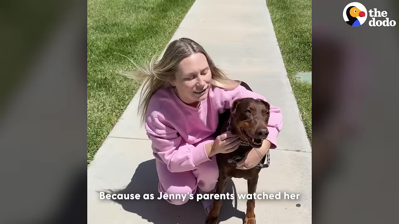 A woman in a pink tracksuit smiles as she kneels on a sidewalk and hugs a brown Doberman dog.