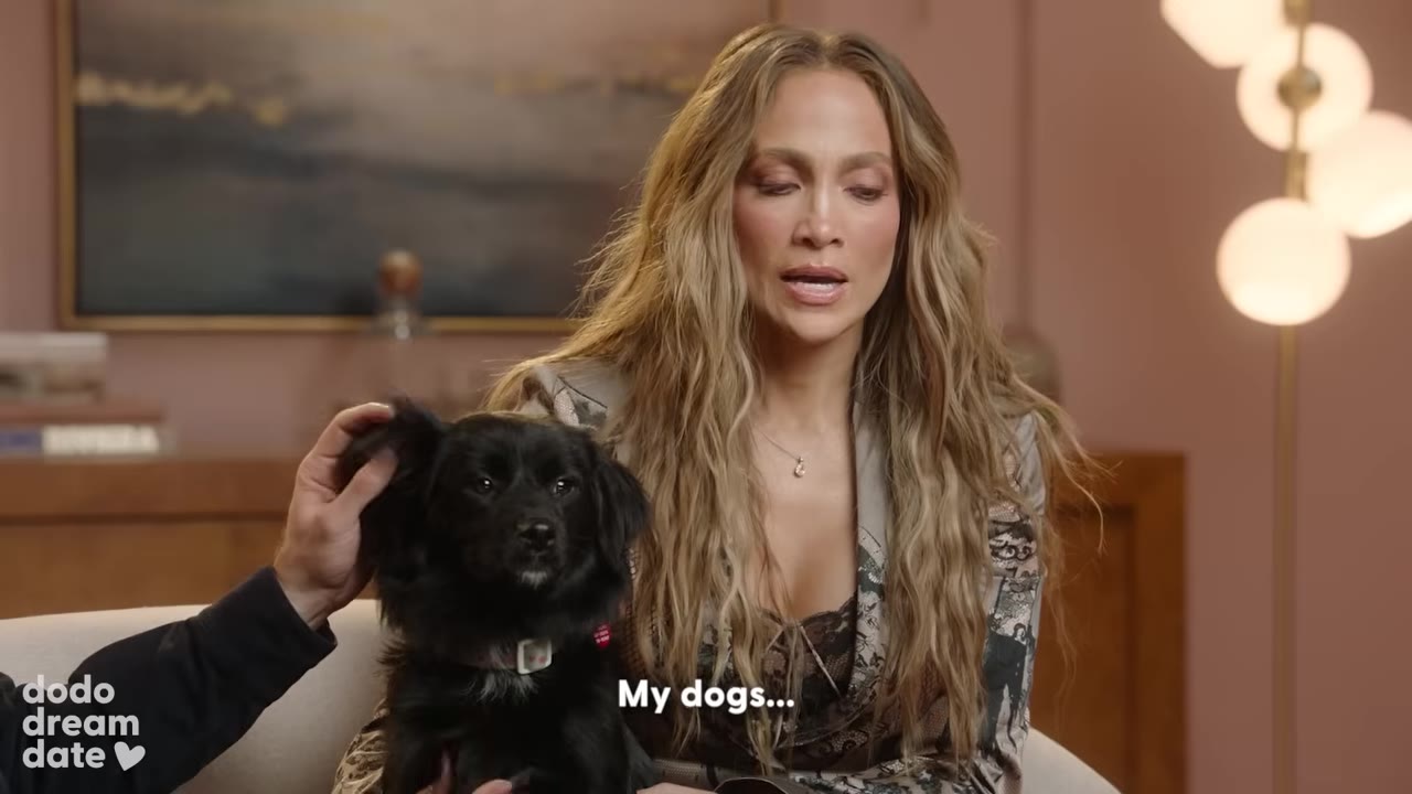 A close-up shot of Jennifer Lopez with long blonde hair, wearing a patterned blazer, holding a small black dog while speaking, with the subtitle 'My dogs...' displayed on screen.
