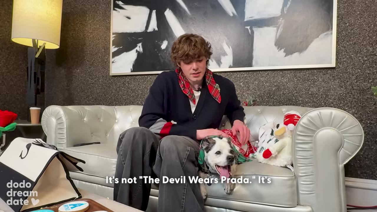 A young man with curly brown hair sits on a white leather couch next to a small white and black dog. The man wears a red plaid bandana around his neck, and the dog wears a matching green patterned bandana.