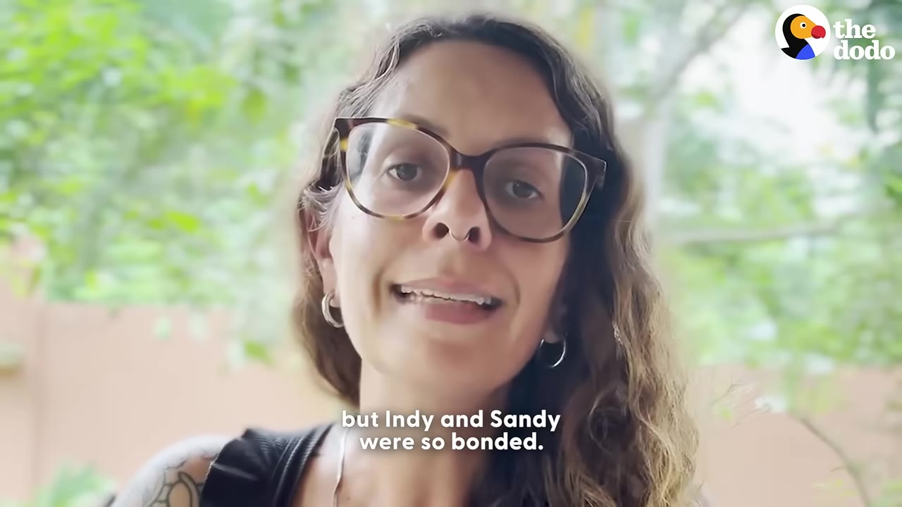 A close-up of Vanessa, a woman with glasses and long curly hair, speaking with a green foliage background.