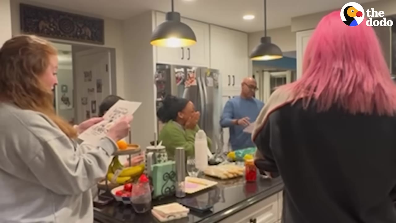 A group of people gathered in a kitchen, with one woman on the left holding and reading a printed flyer while another woman in a green shirt reacts with surprise, covering her mouth with her hands.