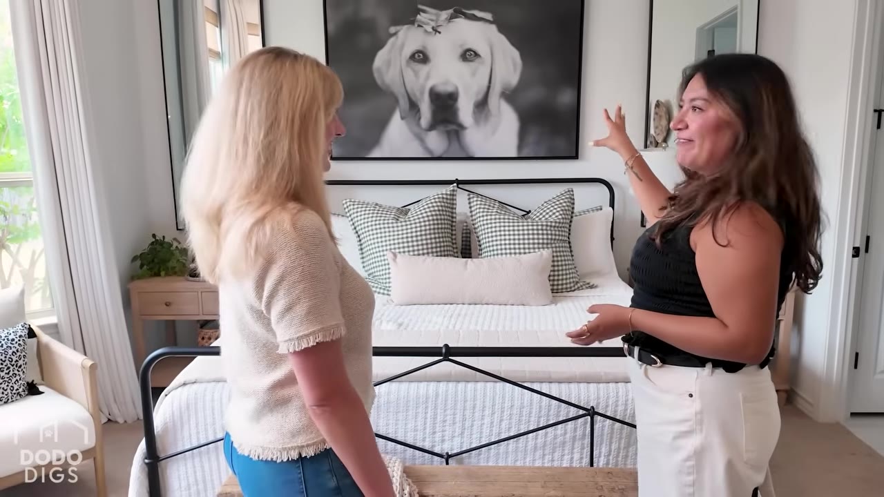 Two women stand in a master bedroom looking at a large black-and-white portrait of a yellow Labrador retriever hanging above the bed.