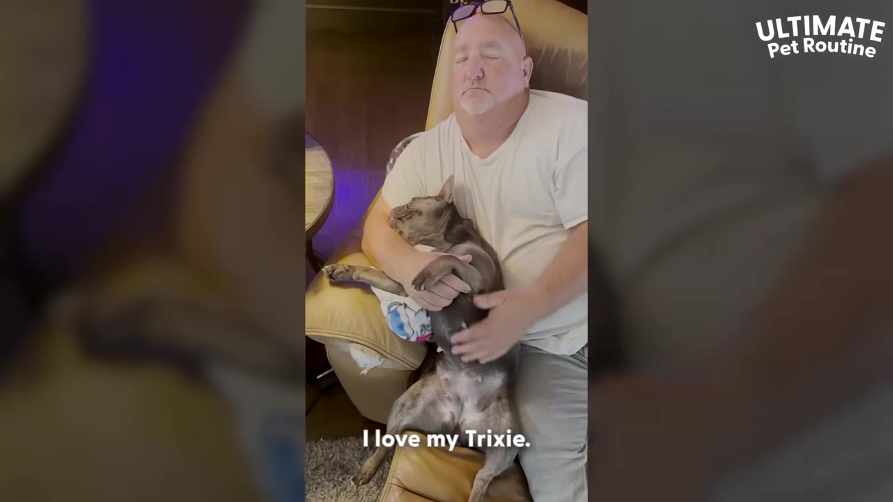 A man sits in a recliner chair with his eyes closed, cradling a grey French bulldog in his lap.